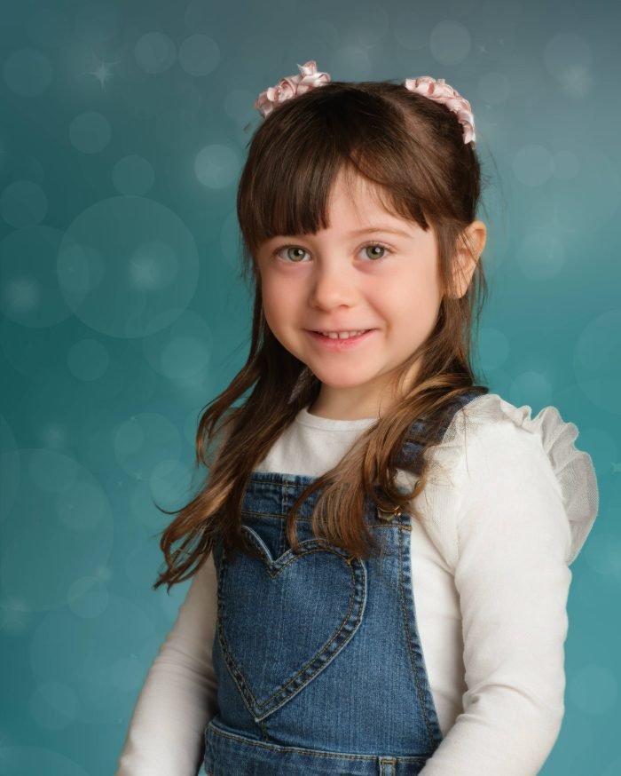Preschool photo day portrait of a girl in denim overalls and a pink headband on a teal backdrop